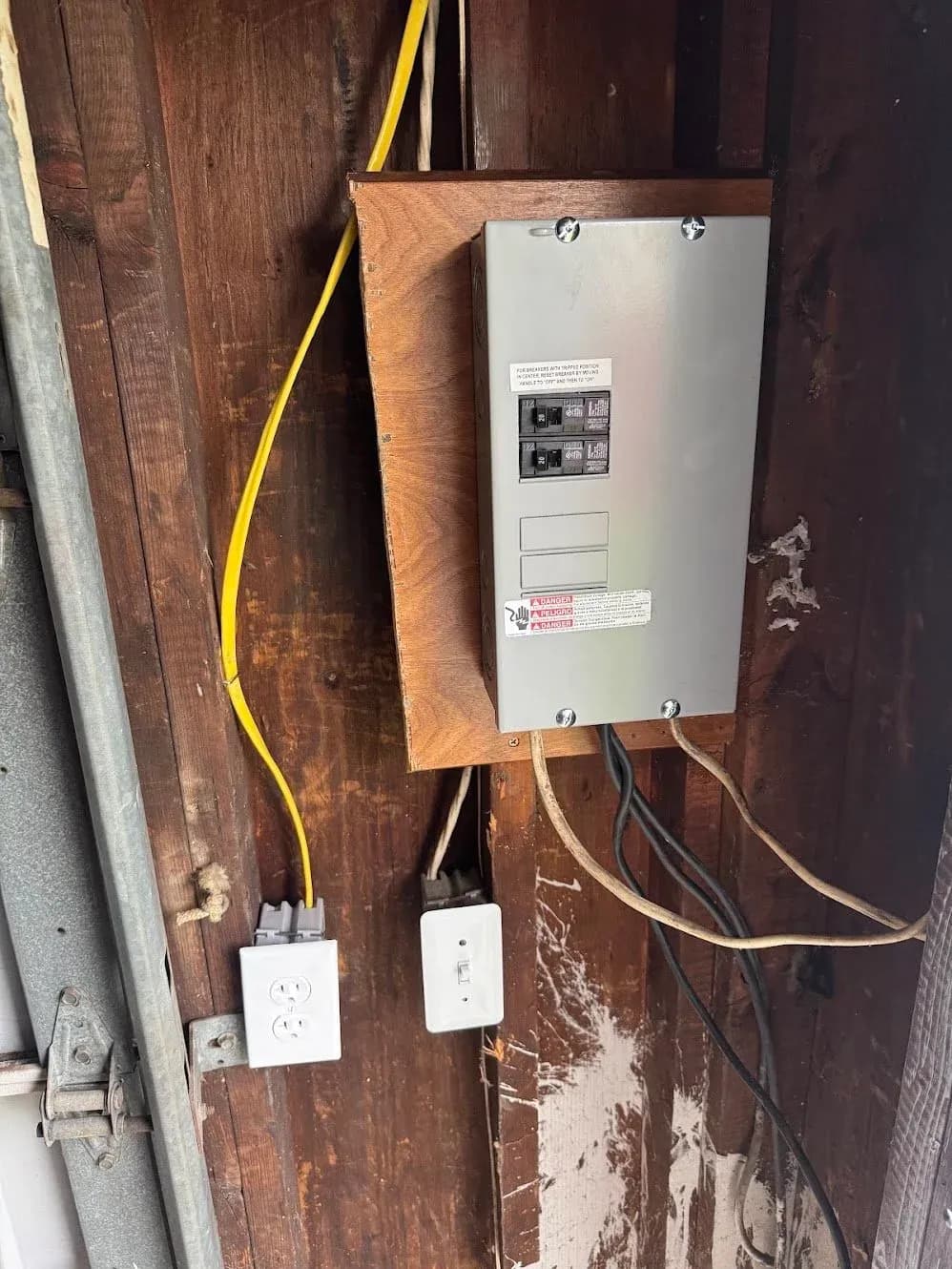 A subpanel mounted on a stained wood wall with a yellow cable run to an outlet and switch below