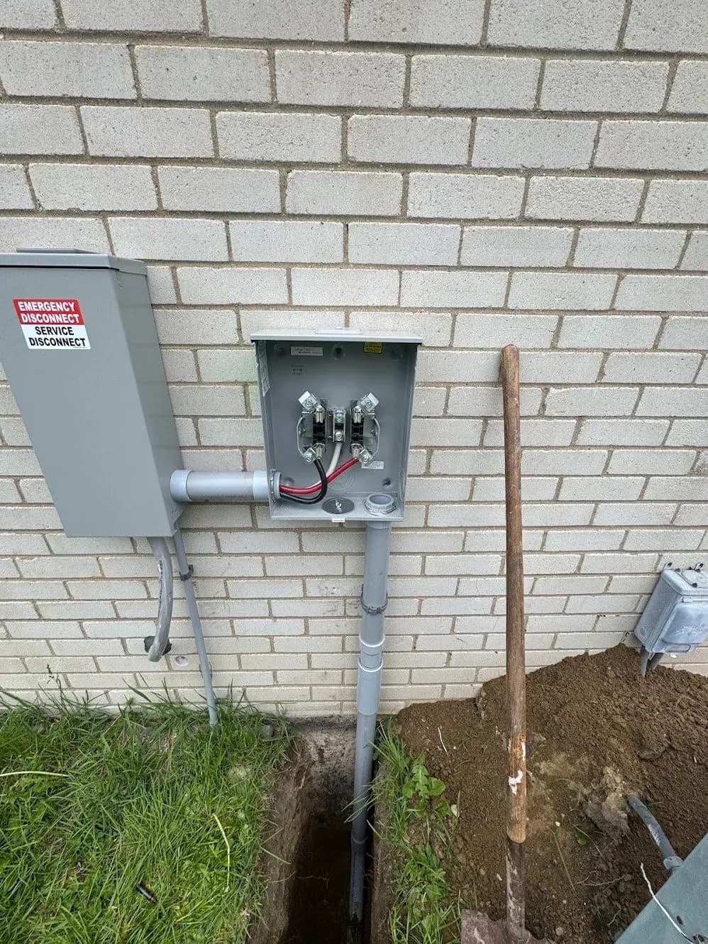 An exterior meter socket and service disconnect on a brick wall above an open trench for underground conduit